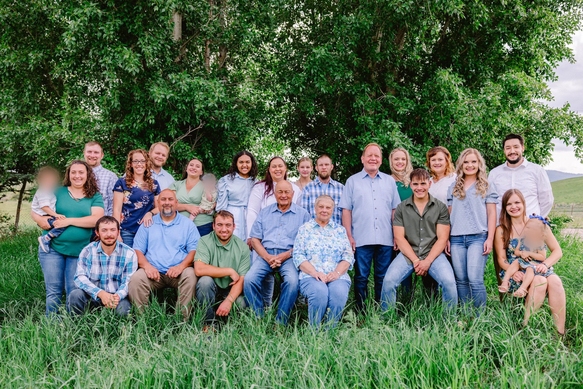 The extended Hokum family in 2023, gathered in tall grass under green trees, with Foalima and his wife seated in the center.